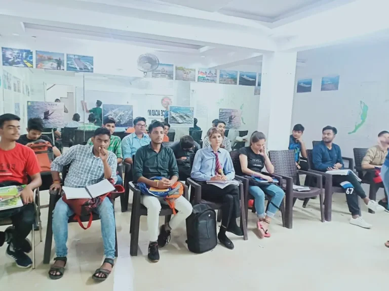 Students seated on chairs in a bright classroom with white walls, some holding notebooks and papers, with posters on the wall behind them