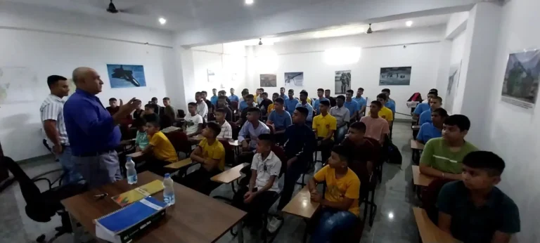 Two instructors, one in a blue shirt, talk to a room of students seated at desks, with framed pictures on the classroom walls