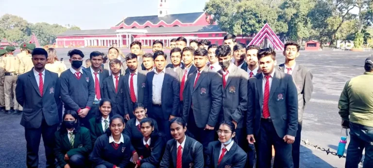 Boys and girls in dark blazers and red ties pose in rows on the parade ground of the Indian Military Academy, with the academy building behind them