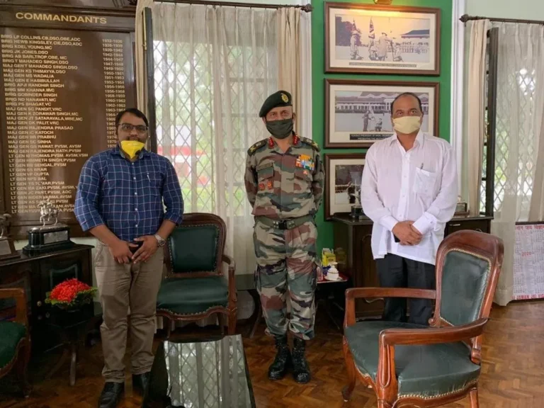 Two men in everyday clothes stand on either side of an Army officer in camouflage uniform and green beret in a room with a board listing the names of commandants