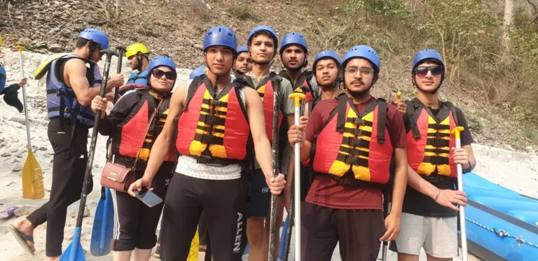 A group of young people in blue helmets and orange life jackets stand on a riverbank holding paddles beside an inflatable raft